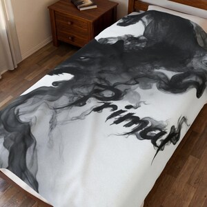 Op de afbeelding: Een witte sprei met een grote, donkergrijze, rookachtige afbeelding die het woord Primal vormt. Het ontwerp bestaat uit wervelende, ijle rookpatronen die zich over de stof uitstrekken en een contrastrijk visueel effect op het oppervlak creëren.