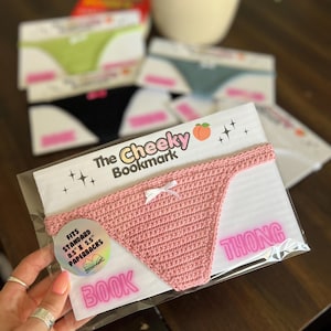May include: A pink crocheted thong bookmark with a white bow, packaged on a white card. The card reads "The Cheeky Bookmark" and "Thong Book." Other bookmarks in green, black, and blue are visible in the background. The bookmark fits standard 8.5" x 5.5" paperbacks.