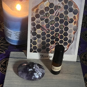 May include: A lit blue candle with a white and purple wrapped top sits next to a wooden tray holding a polished purple stone, a small brown bottle, and a tarot card with a honeycomb and bee illustration. The card reads "Nest of".