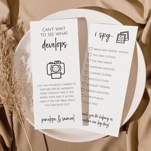 May include: Two white cards on a white plate with text and illustrations. One card reads "Can't wait to see what develops" with a camera graphic. The other card says "I spy" with a list of wedding activities. The cards are on a beige cloth.