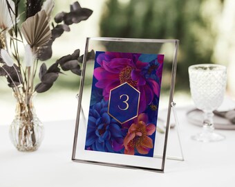 May include: A clear glass table number holder with a floral design. The card features a vibrant floral arrangement in shades of purple, blue, and orange, with the number "3" in a gold hexagon. A vase of dried flowers and a water glass are in the background.