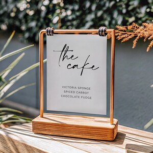 May include: A wooden table sign with a copper frame, displaying the text "the cake" in elegant script. Below, the sign lists cake flavors: Victoria Sponge, Spiced Carrot, and Chocolate Fudge. The sign is set on a wooden base.