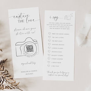 May include: Two white wedding cards with black text. One card says "Capture the Love" and includes a camera graphic with a QR code. The other card says "I Spy..." with a list of wedding moments. A white ribbon and dried plant are also in the image.