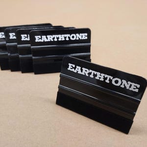 Puede incluir: Varias espátulas negras con la palabra "EARTHTONE" en letras blancas. Las espátulas tienen un mango negro y un borde de fieltro negro. Están dispuestas sobre una superficie marrón claro.
