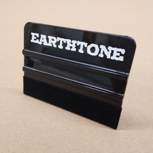Puede incluir: Una espátula negra con el texto blanco "EARTH TONE" impreso en la parte superior. La espátula tiene un borde de fieltro negro. La herramienta está diseñada para aplicar gráficos y calcomanías de vinilo.