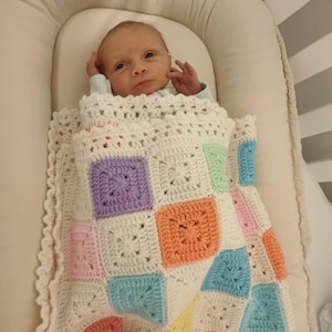 May include: A baby wrapped in a handmade, colourful crochet blanket. The blanket features square patterns in pastel colours like pink, blue, orange, and purple, set against a white background. The baby is in a white bassinet.