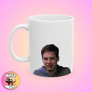 Può includere: Tazza in ceramica bianca con l'immagine di un uomo che piange. L'uomo ha i capelli castani e indossa una felpa grigia.