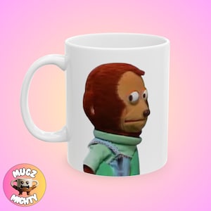 May include: White ceramic coffee mug with a brown and green monkey puppet with one eye looking directly at the camera. The monkey is wearing a green shirt and blue overalls.