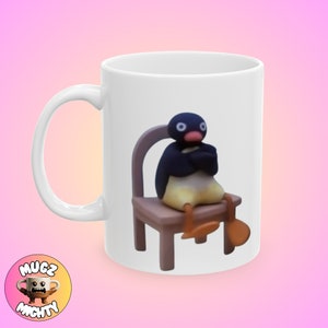 May include: White ceramic mug featuring a printed image of a cartoon penguin sitting on a wooden chair with a confused expression.