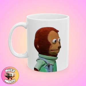 May include: White ceramic coffee mug with a brown and red monkey puppet with big eyes looking to the right. The monkey is wearing a green and blue shirt.