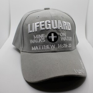 May include: A gray baseball cap with the word "LIFEGUARD" embroidered in white. Below the word "LIFEGUARD" is the text "MINE WALKS ON WATER" and "MATTHEW 14:25-33". A black circle with a white cross is also embroidered on the cap.