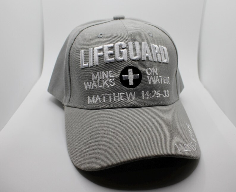 May include: A gray baseball cap with the word "LIFEGUARD" embroidered in white. Below the word "LIFEGUARD" is the text "MINE WALKS ON WATER" and "MATTHEW 14:25-33". A black circle with a white cross is also embroidered on the cap.