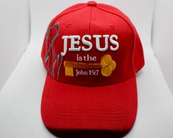 May include: A red baseball cap with the words "JESUS is the" and "John 15:7" embroidered in white and gold. The cap has a curved brim and a button on top. A gray design is on the side.