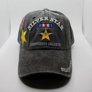 May include: A gray baseball cap with embroidered text and star designs. The cap features the words "SILVER STAR" and "CONSPICUOUS GALLANTRY" in white thread. A gold star and a red, white, and blue emblem are also embroidered on the cap.