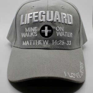 May include: A gray baseball cap with white embroidered text that reads "LIFEGUARD" above "MINE WALKS ON WATER" and "MATTHEW 14:25-33". A black cross is centered. The brim has "I LOVE JESUS" embroidered.