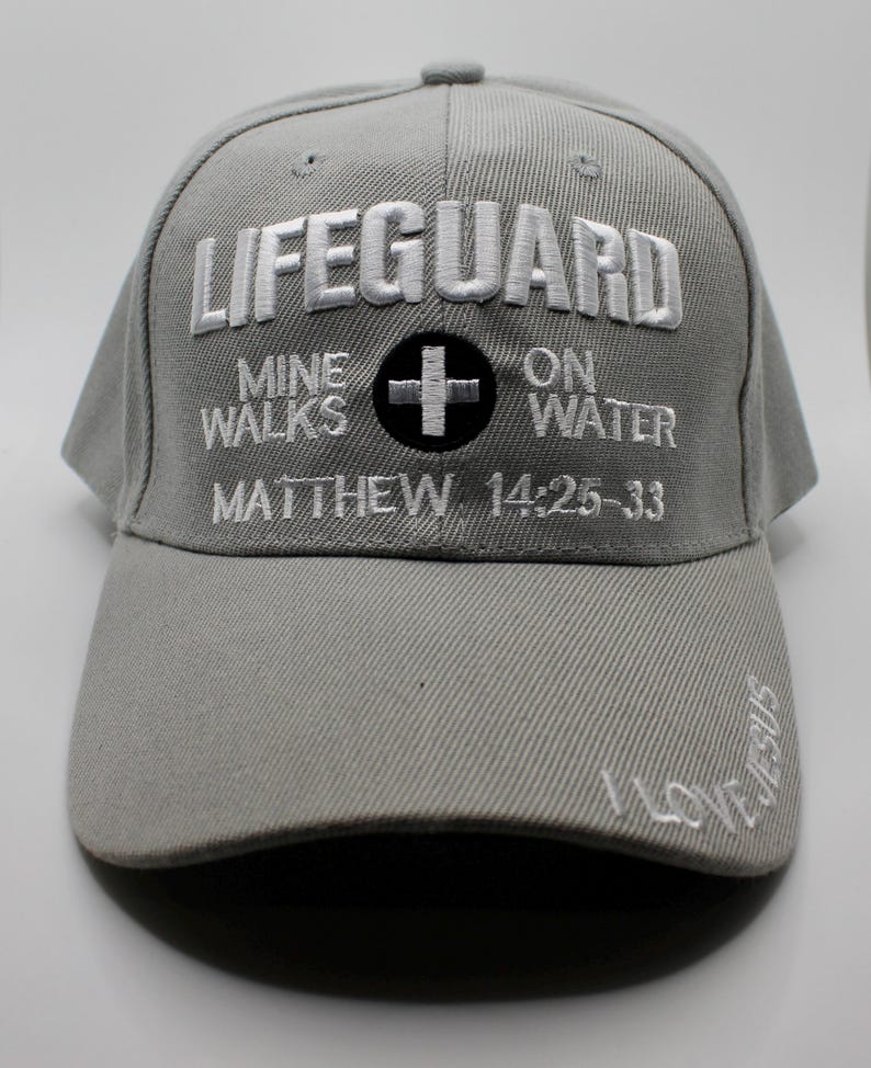 May include: A gray baseball cap with white embroidered text that reads "LIFEGUARD" above "MINE WALKS ON WATER" and "MATTHEW 14:25-33". A black cross is centered. The brim has "I LOVE JESUS" embroidered.