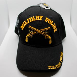 May include: Black baseball cap with a gold embroidered military police logo featuring crossed pistols and the words "Military Police".