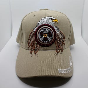 May include: A tan baseball cap featuring an embroidered design of a white eagle with a yellow beak positioned above a circular emblem with brown and white patterns. The cap includes brown feather accents and the text NATIVE PRIDE in white embroidery.