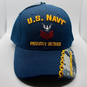 May include: A navy blue baseball cap with the text "U.S. NAVY" embroidered in gold. The cap features a white eagle emblem and red chevrons. The phrase "PROUDLY SERVED" is also embroidered in gold.