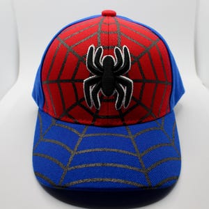 Puede incluir: Gorra de béisbol con diseño de Spider-Man. La gorra tiene una parte superior roja con un patrón de telaraña negra y un emblema de araña negra. La visera y los laterales son azules.