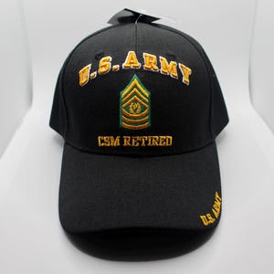 Puede incluir: Gorra de béisbol negra con bordado dorado. La gorra dice "U.S. ARMY" y "CSM RETIRED". Un distintivo de rango dorado y verde está bordado en la parte delantera.