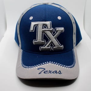 Pode incluir: Um boné de beisebol azul e cinza com uma aba branca costurada. O boné tem um logotipo "TX" bordado em cinza com a palavra "Texas" bordada em branco na aba.