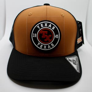 May include: A brown and black baseball cap with a mesh back. The front of the cap features a circular patch with the word "TEXAS" and the letters "Tx" in red. The brim is black.