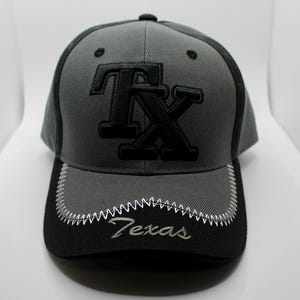May include: A grey and black baseball cap featuring the letters "TX" in black embroidery. The black brim has "Texas" embroidered in white, with a white zigzag stitch detail.