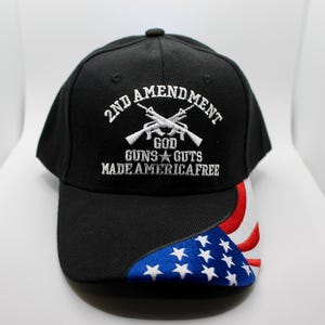 Könnte beinhalten: Schwarze Baseballkappe mit weiß gesticktem Text: "2ND AMENDMENT GOD GUNS & GUTS MADE AMERICA FREE." Der Schirm der Kappe zeigt ein Design der amerikanischen Flagge mit roten, weißen und blauen Farben.