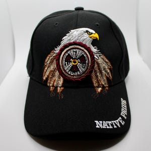 Könnte beinhalten: Schwarze Baseballkappe mit einem gestickten Adlerdesign. Der Adler hat weiße Federn, einen gelben Schnabel und ist vor einem indianisch inspirierten Emblem platziert. Die Worte "NATIVE PRIDE" sind auf der Seite gestickt.