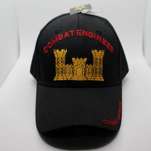 May include: Black baseball cap with "COMBAT ENGINEER" embroidered in red. The cap features a gold emblem of a castle. The text "COMBAT ENGINEER" is also embroidered on the bill.