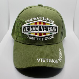 Puede incluir: Una gorra de béisbol verde con un parche bordado blanco que dice "Vietnam Veteran" y una franja amarilla, roja y verde. La gorra también tiene el texto "Time Was Served" y "Time To Honor" bordado.