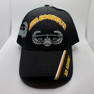 May include: Black baseball cap with gold embroidered "AIR ASSAULT" text. It features a white and grey helicopter and wing emblem. The cap has a curved peak and a white, gold, and black stripe on the side.