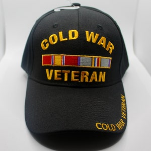 May include: A black baseball cap featuring the embroidered yellow text COLD WAR VETERAN and a colorful ribbon bar graphic on the front. The words COLD WAR VETERAN are also embroidered in yellow on the right side of the curved brim of the cap.