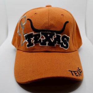 May include: Orange baseball cap featuring embroidered "TEXAS" lettering and a longhorn graphic. The cap has a curved peak and a small embroidered "TEXAS" on the peak.