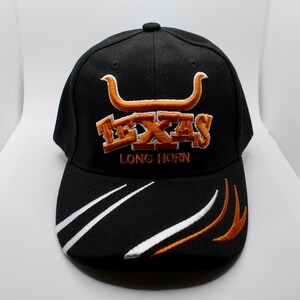 Può includere: Cappellino da baseball nero con ricami arancioni e bianchi. Il cappellino presenta la scritta "TEXAS" e un simbolo di longhorn in arancione, con "LONG HORN" sotto. Linee curve bianche e arancioni adornano la visiera.