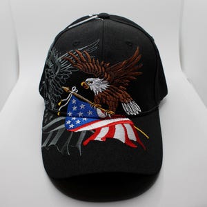 Può includere: Cappellino da baseball nero con un design ricamato raffigurante un'aquila marrone e bianca che tiene una bandiera americana. Il cappellino ha una visiera curva e una rappresentazione dettagliata della bandiera americana.