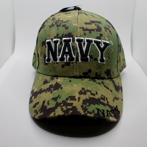 以下が含まれることがあります： 緑と黒の迷彩柄のベースボールキャップで、前面に黒で「NAVY」と刺繍されています。