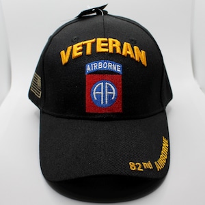 以下が含まれることがあります： 黒い野球帽。金色の刺繍で「VETERAN」の文字。つばには「AIRBORNE」のパッチと「82nd AIRBORNE」の文字。側面に小さなアメリカ国旗のパッチが付いています。