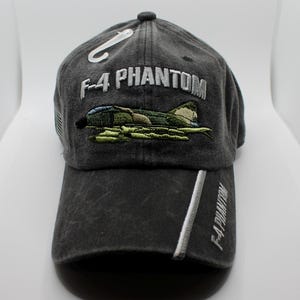 May include: A charcoal gray baseball cap with the text "F-4 PHANTOM" embroidered in white. The cap features an embroidered image of a military aircraft in green, brown, and blue. A white stripe runs down the front of the bill.