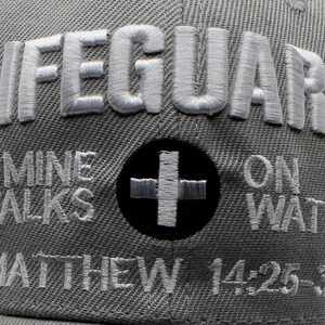 May include: Gray baseball cap with the word "LIFEGUARD" embroidered in white. Below, the text "MINE WALKS ON WATER" and "MATTHEW 14:25-33" are also embroidered. A black circle with a white cross is centered.