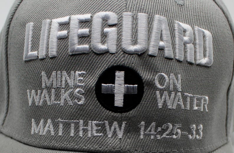 May include: Gray baseball cap with the word "LIFEGUARD" embroidered in white. Below, the text "MINE WALKS ON WATER" and "MATTHEW 14:25-33" are also embroidered. A black circle with a white cross is centered.