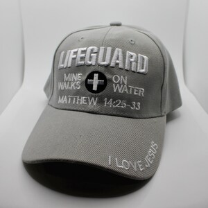 May include: A gray baseball cap with the word "LIFEGUARD" embroidered in white across the front. Below the word "LIFEGUARD" is the text "MINE WALKS ON WATER" and "MATTHEW 14:25-33". The brim of the cap has the words "I LOVE JESUS".