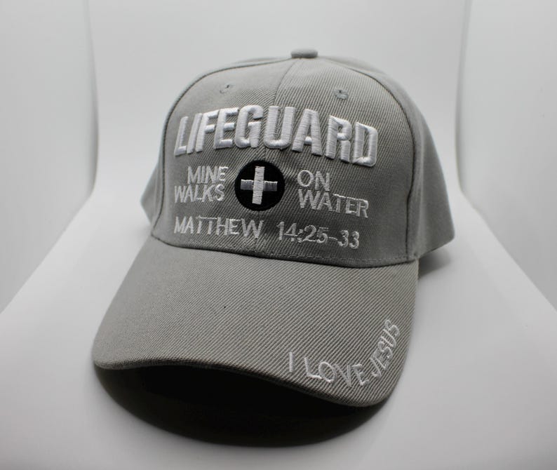 May include: A gray baseball cap with the word "LIFEGUARD" embroidered in white across the front. Below the word "LIFEGUARD" is the text "MINE WALKS ON WATER" and "MATTHEW 14:25-33". The brim of the cap has the words "I LOVE JESUS".