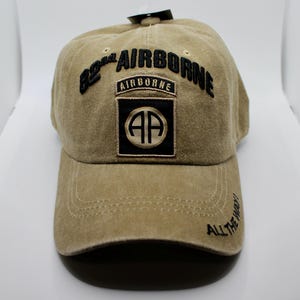 May include: A tan baseball cap with the text "82nd AIRBORNE" embroidered in black. The cap features a patch with "AIRBORNE" and "AP" in a black and gold design. The phrase "ALL THE WAY!" is embroidered on the brim.