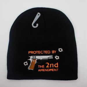 May include: Black beanie with the embroidered text "PROTECTED BY THE 2ND AMENDMENT" in orange. The design includes a pistol and bullet holes. A silver plastic tag is attached to the top of the beanie.