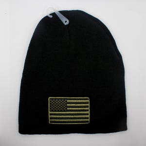 May include: Black knitted beanie hat featuring an embroidered American flag patch in olive green and tan. The hat has a simple design and a hang tag attached at the top.