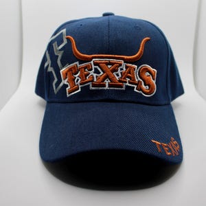 Könnte beinhalten: Eine marineblaue Baseballkappe mit dem in Orange und weißem Umriss gestickten Wort TEXAS. Über dem Text befindet sich ein orangefarbenes Longhorn-Stier-Logo. Auf der linken Seite ist ein silberner Umriss des Bundesstaates Texas und auf dem Schirm ist das Wort TEXAS in Orange gestickt.