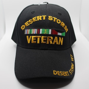 Puede incluir: Gorra de béisbol negra con bordado dorado que dice "Desert Storm Veteran" y presenta una fila de cintas militares y una bandera.