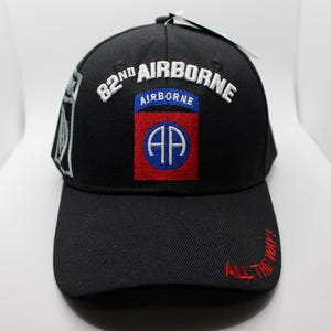 May include: Black baseball cap with the text "82nd AIRBORNE" in white embroidery. The cap features a patch with "AP" in blue and a red border. The brim has the embroidered text "ALL THE WAY!" in red.